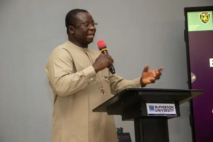 Osuolale A. Festus speaking into a microphone at a lectern marked McPherson University.